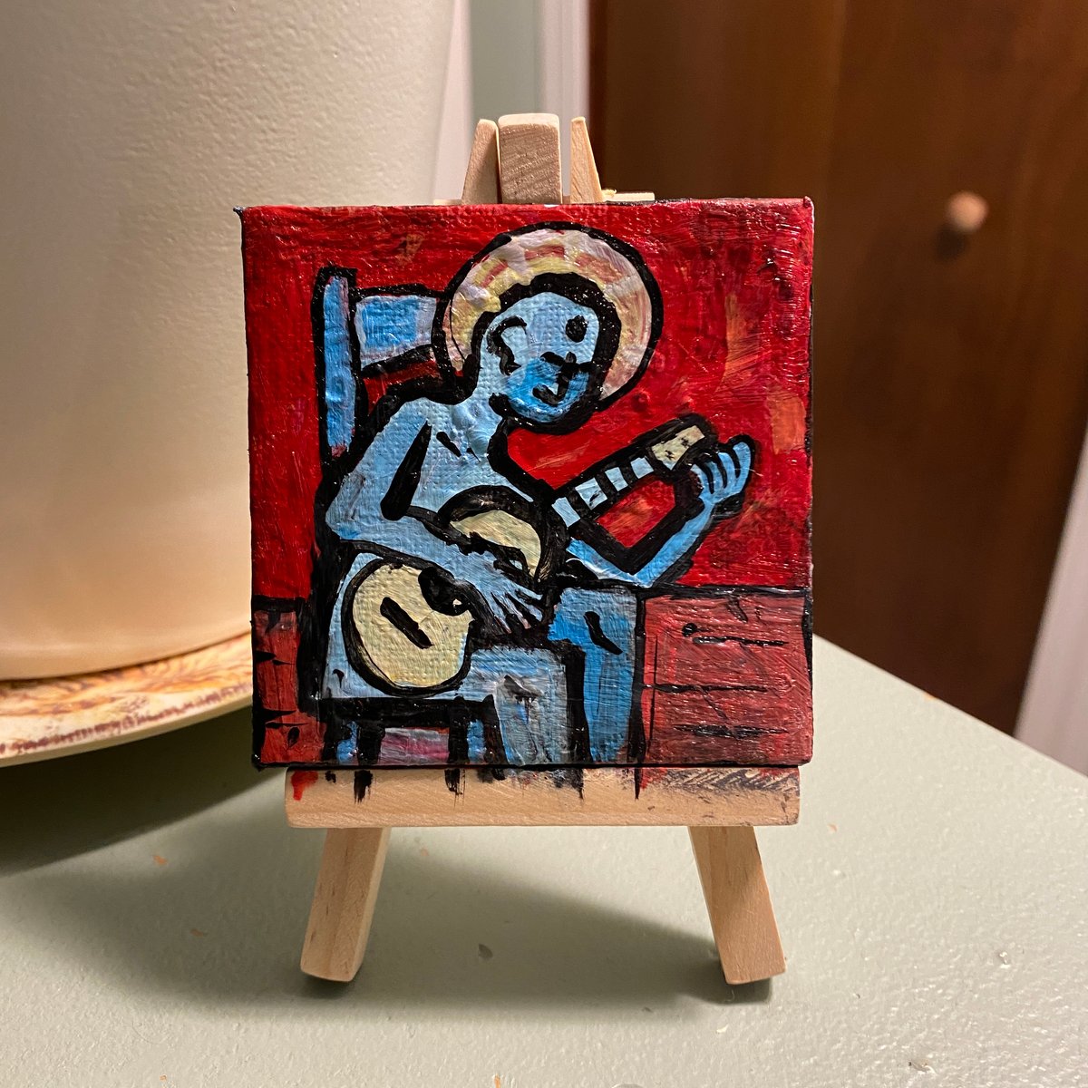 Image of haloed blue being tuning a guitar while sitting on blue chair in a red room with a wooden floor